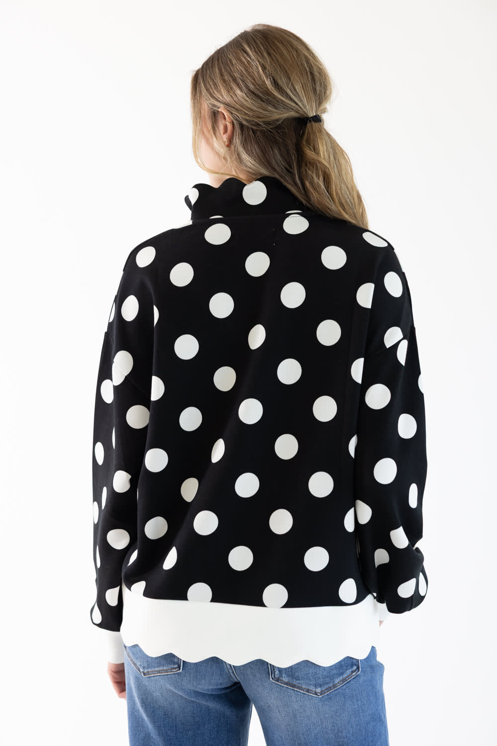 A woman viewed from the back wearing a black long-sleeve top with large white polka dots, layered over a white scalloped hem shirt and paired with blue jeans, against a plain white background.