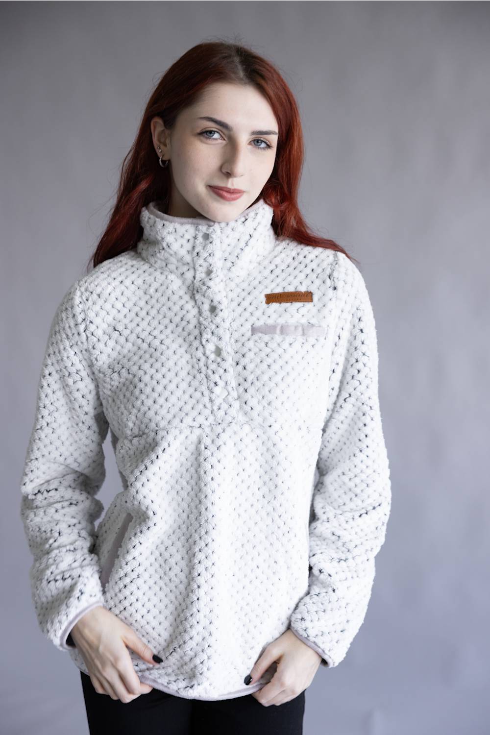 A woman with red hair wearing a white textured fleece pullover jacket with a high collar and a small brown chest patch, posed against a neutral gray studio background.