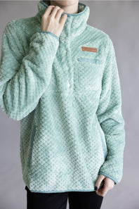 A person wearing a light mint green textured fleece pullover jacket with a stand collar and a small brown rectangular logo patch on the chest, photographed against a plain gray studio background.