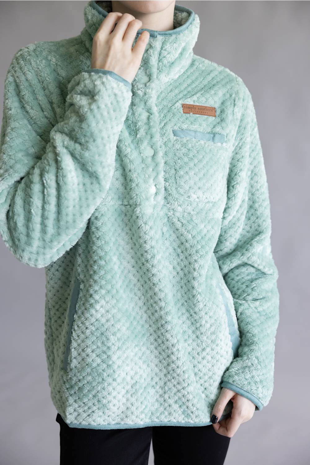 A person wearing a light mint green textured fleece pullover jacket with a stand collar and a small brown rectangular logo patch on the chest, photographed against a plain gray studio background.
