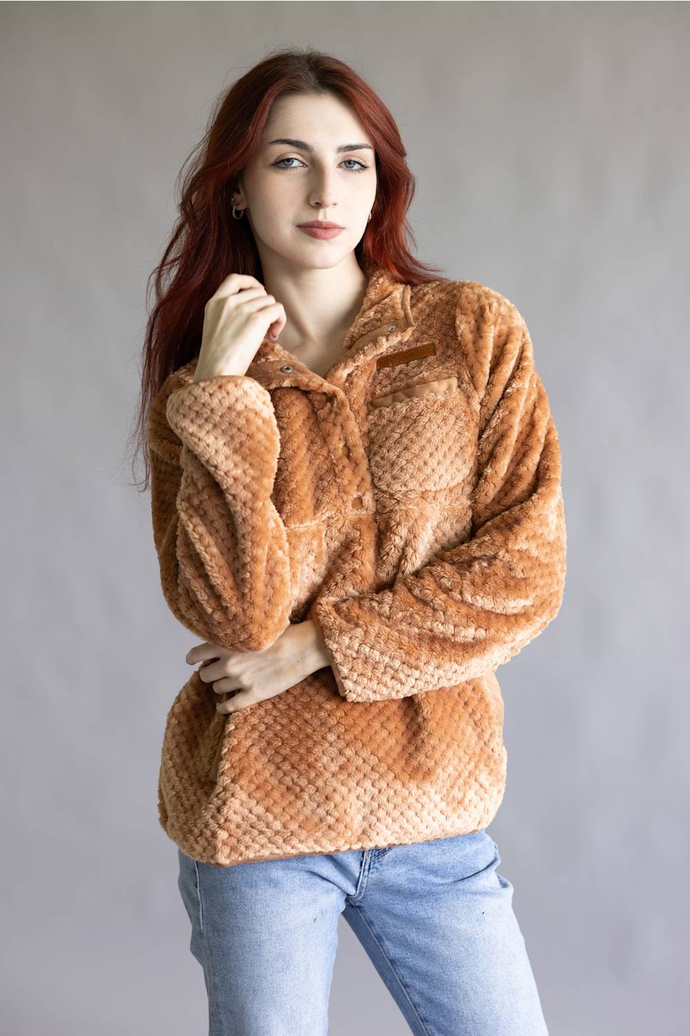 A woman with red hair wearing a textured, long-sleeve brown faux fur jacket and light blue jeans, standing against a plain gray studio background.