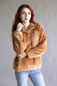 A woman with red hair wearing a textured, long-sleeve brown faux fur jacket and light blue jeans, standing against a plain gray studio background.