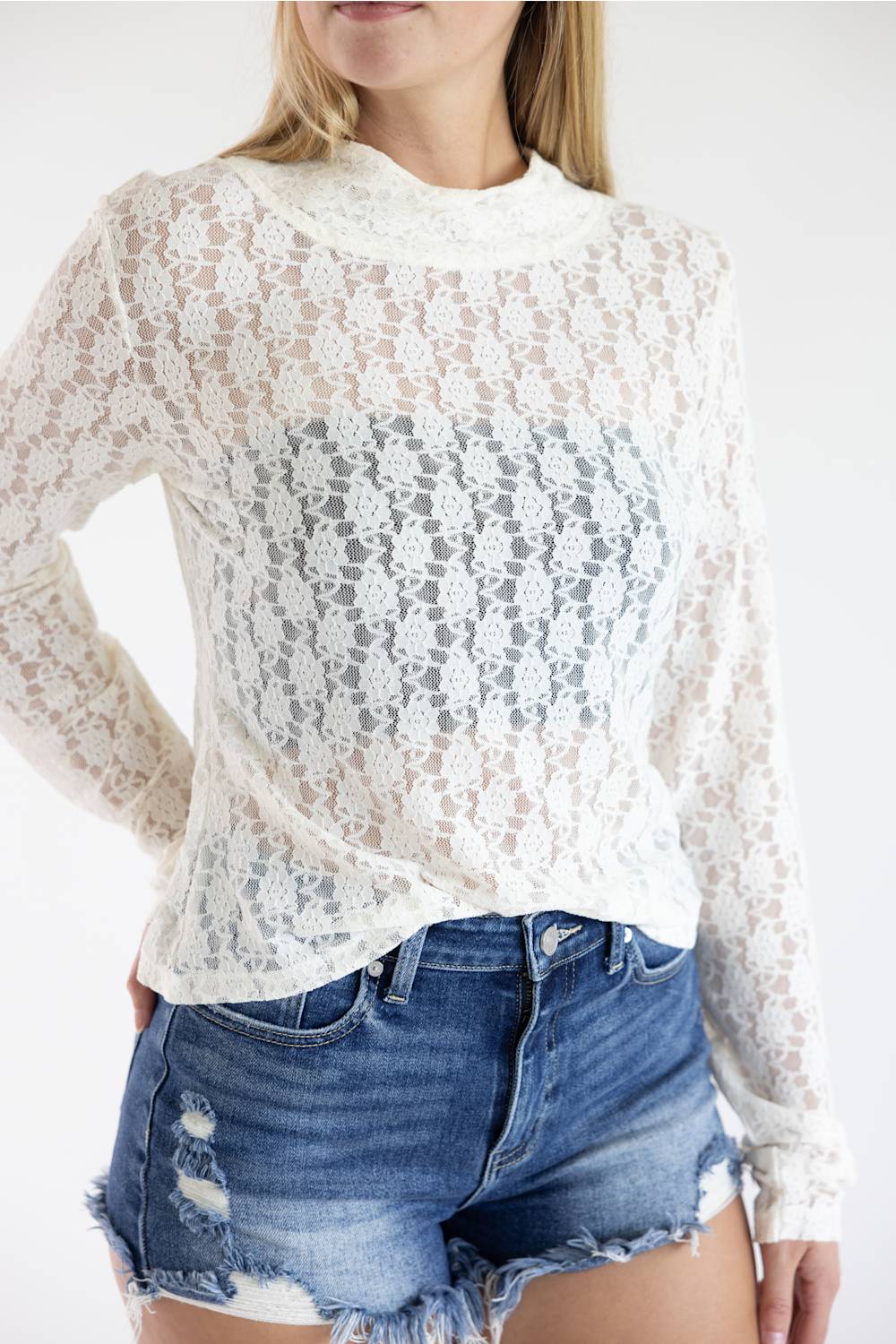 A woman wearing a white long-sleeve lace mock-neck top and distressed denim shorts against a plain white background.