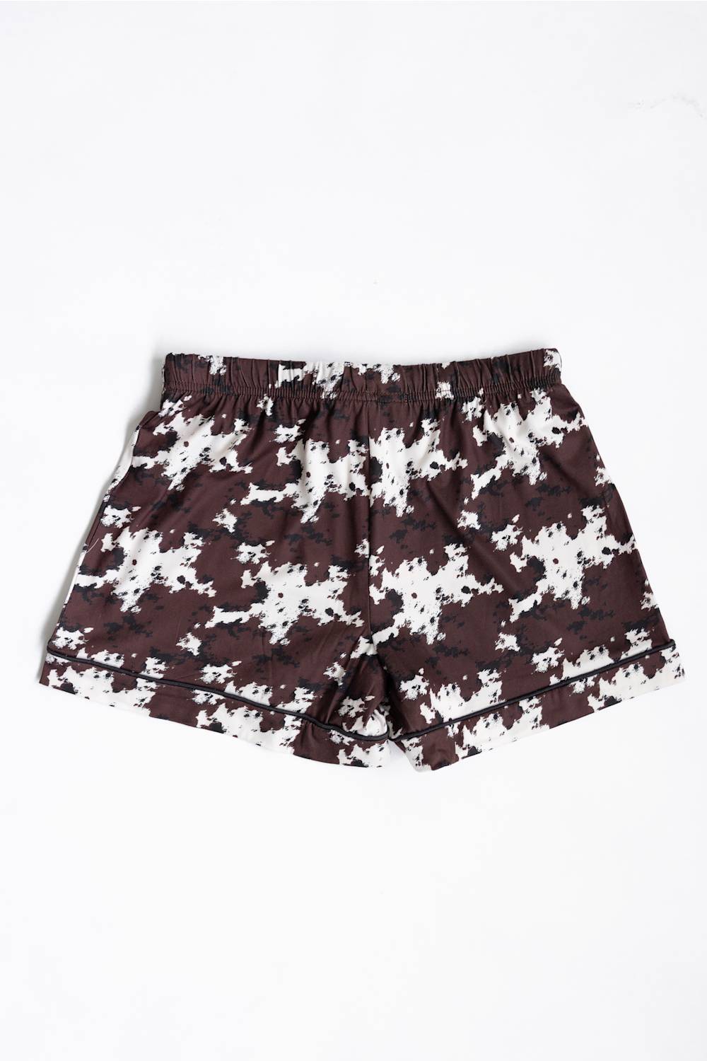 A pair of youth girls shorts featuring a dark brown and white cow pattern, with a black drawstring at the elastic waistband, laid flat against a white background.