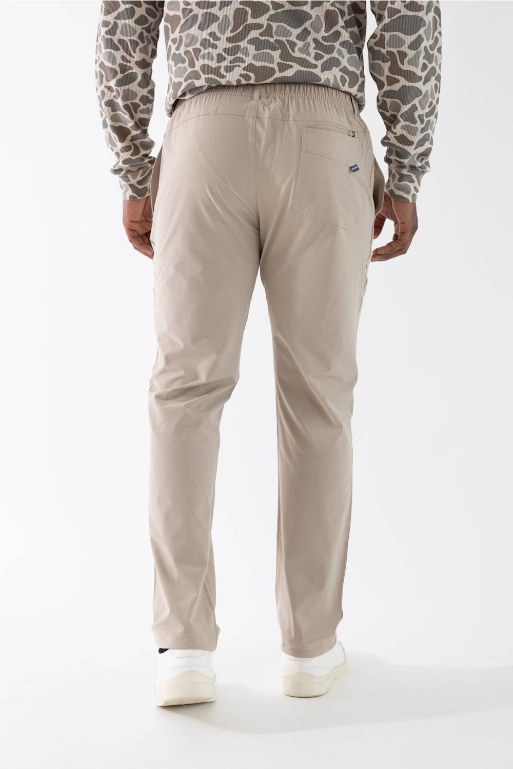 A person shown from the back wearing light beige straight-leg trousers, a beige and gray patterned long-sleeve shirt, and white sneakers against a plain white studio background.