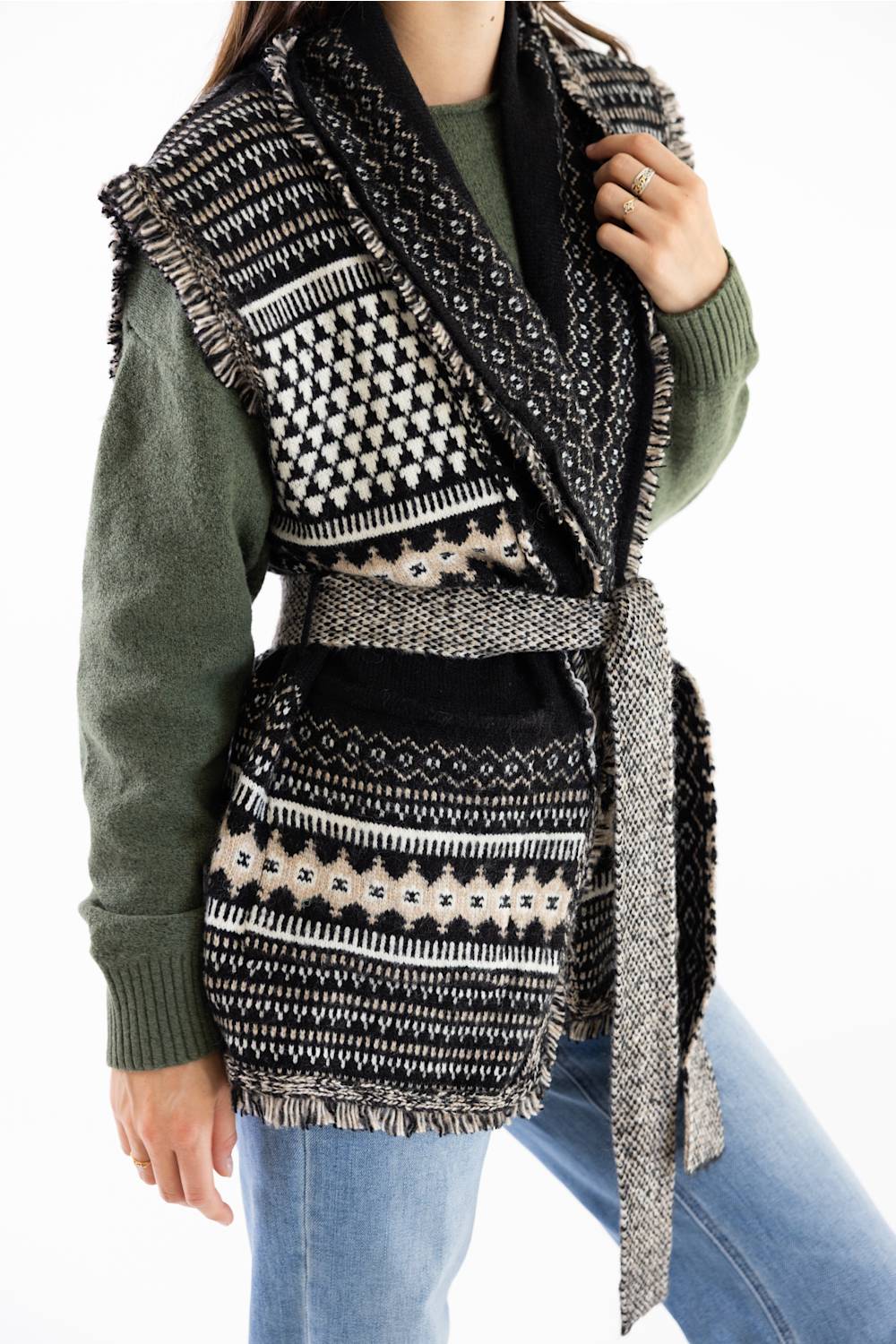 A person wearing a black and cream patterned knit belted vest over an olive green sweater and blue jeans, shown against a plain white background.