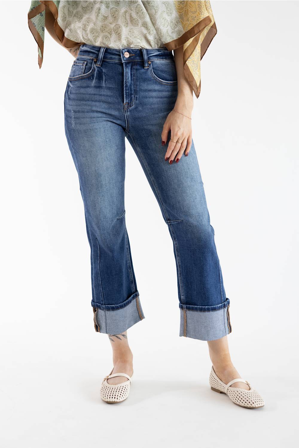 A model wearing a pair of medium-wash blue cropped jeans with rolled cuffs, paired with a patterned blouse and beige flat shoes, shot against a plain white studio background.