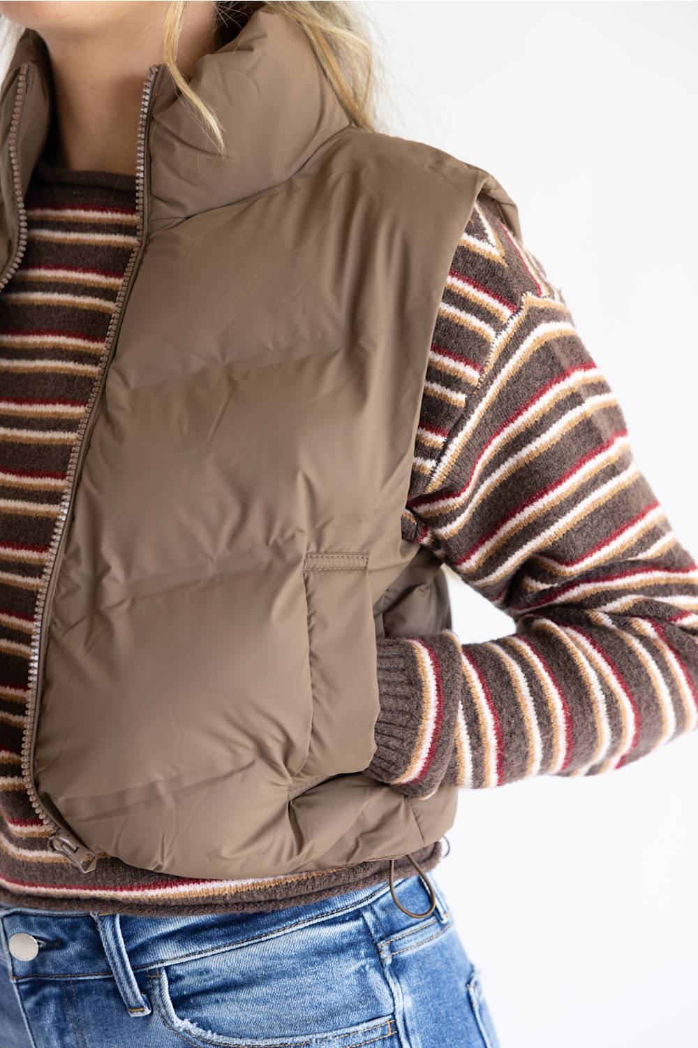 A person wearing a taupe brown puffer vest over a brown, red, and cream striped knit sweater, paired with blue jeans, shown against a plain white background.