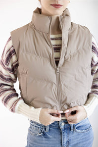 A person wearing a sleeveless beige puffer vest over a cream and brown striped knit sweater, paired with blue jeans, against a plain white background.