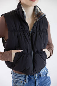 A woman wearing a black quilted puffer vest over a brown long-sleeve top and blue jeans, shown from the neck down against a plain light background.