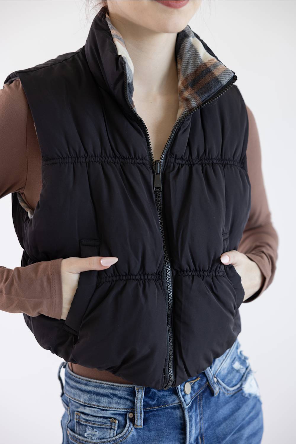 A woman wearing a black quilted puffer vest over a brown long-sleeve top and blue jeans, shown from the neck down against a plain light background.