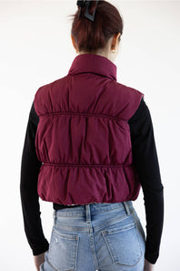 A woman shown from behind wearing a sleeveless quilted puffer vest in deep burgundy over a black long-sleeve top and light blue jeans, against a plain white background.