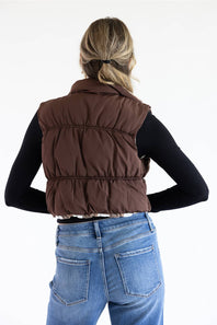 A woman shown from the back wearing a brown puffer vest over a black long-sleeve top and blue jeans, against a plain white studio background.