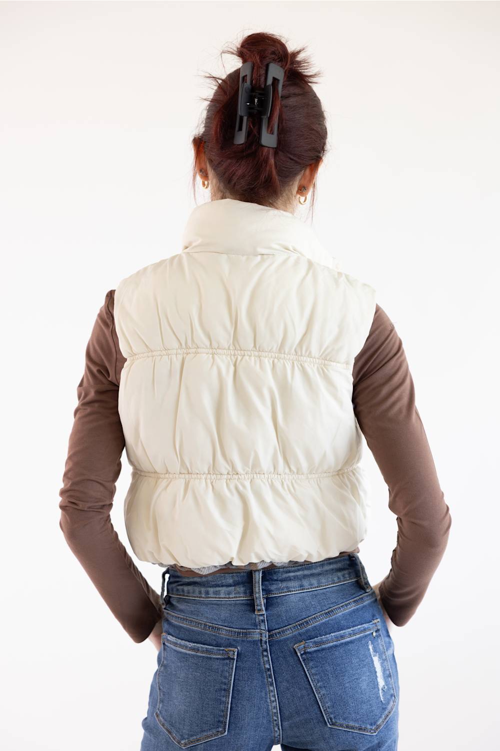 A woman shown from behind wearing a cream-colored puffer vest over a brown long-sleeve shirt and blue jeans, with her hair in a high bun and a black claw clip.