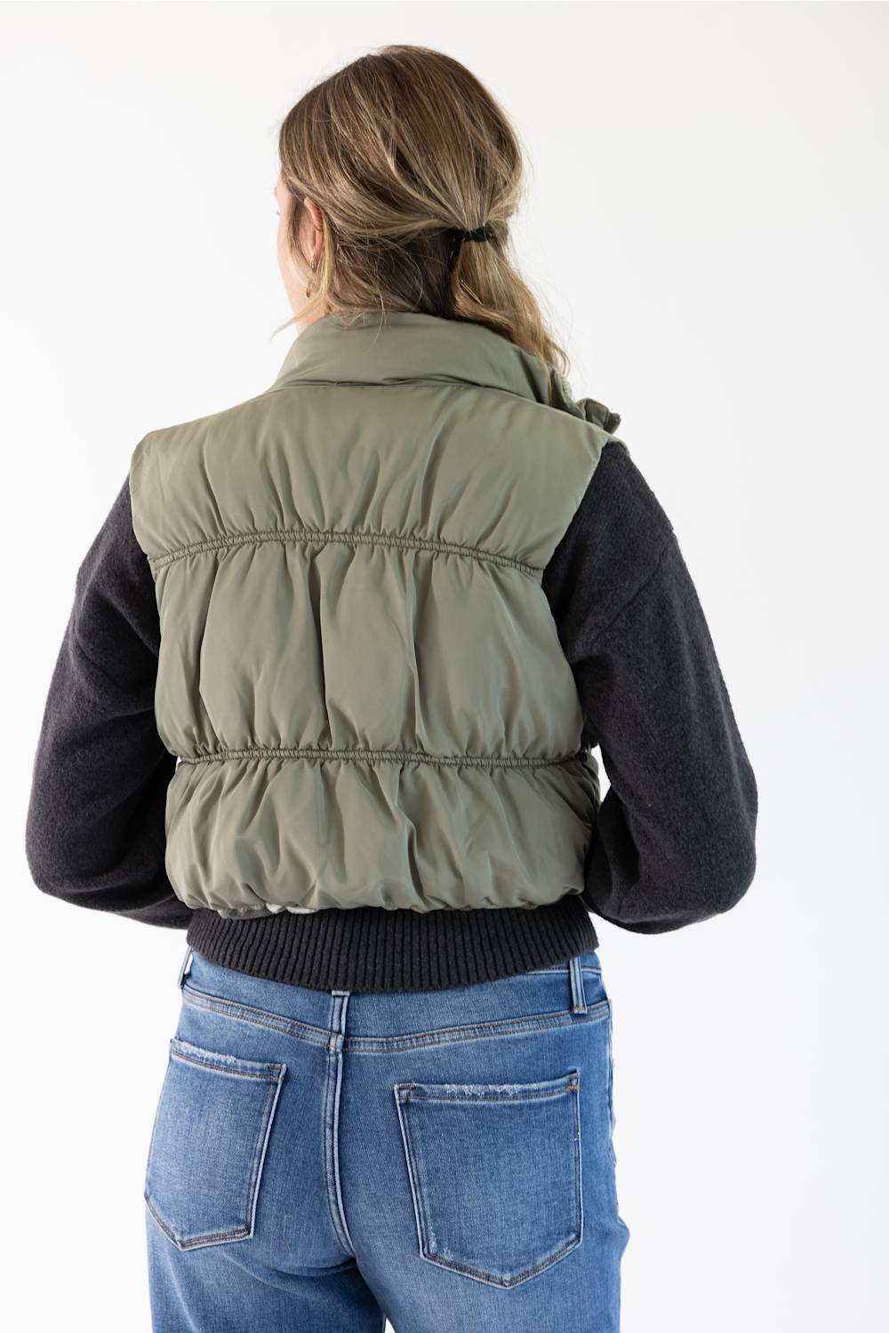 A woman viewed from behind wearing an olive green puffer vest over a black long-sleeve top and blue jeans, against a plain white studio background.