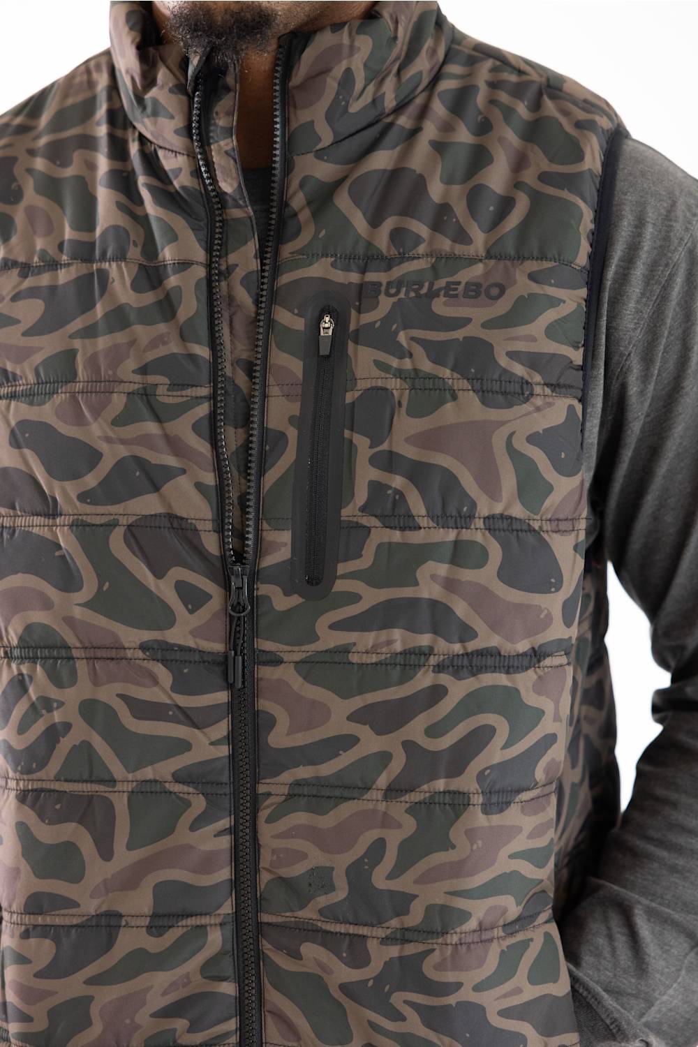 A close-up of a camouflage-patterned puffer vest worn over a dark gray long-sleeve shirt. The vest features a full front zipper, a vertical zippered chest pocket, and the brand name  printed on the left chest.