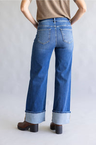 Rear view of a person wearing blue wide-leg cuffed jeans with a tan top and brown block-heel boots against a light gray studio background.