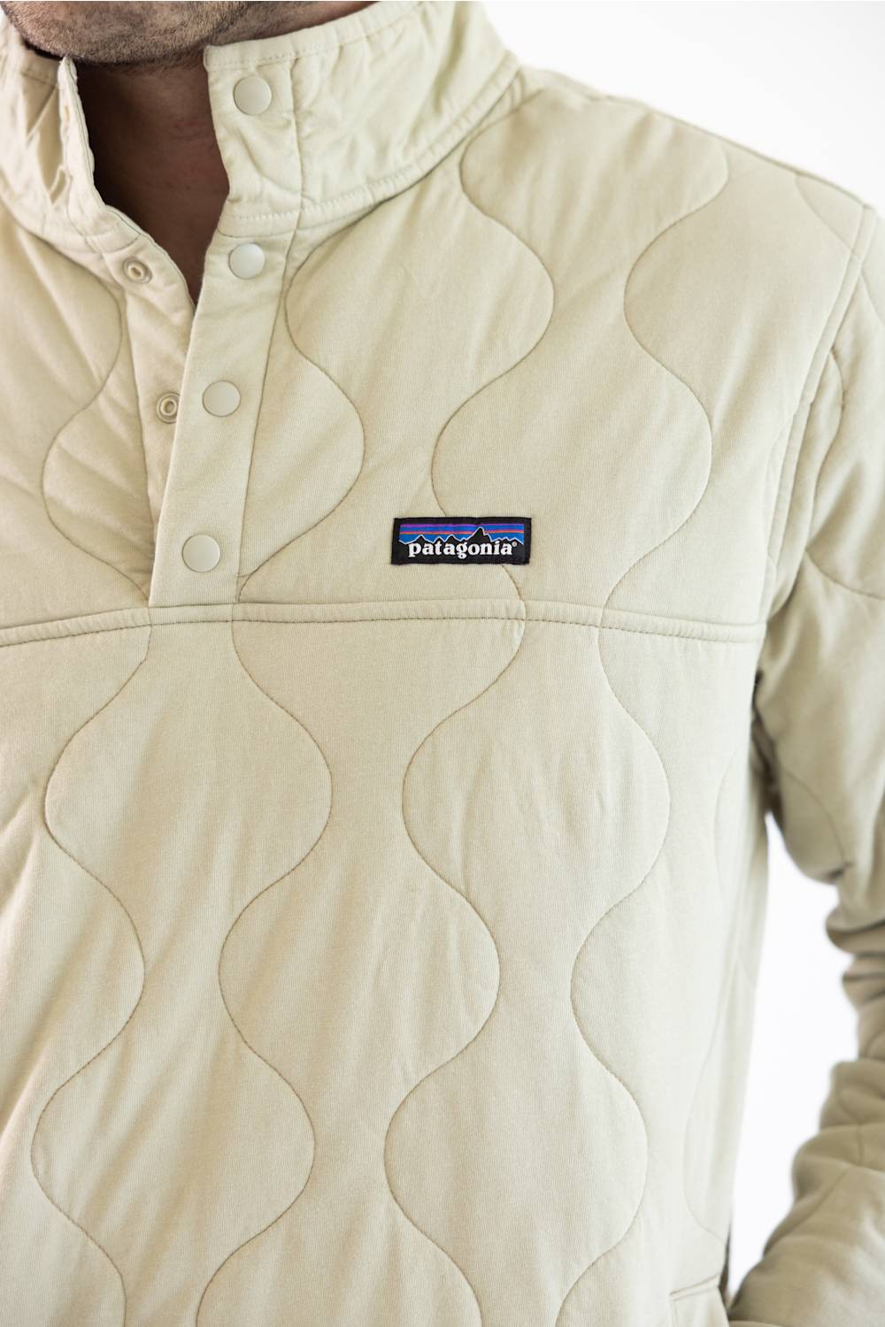 A close up of a man wearing an off white Patagonia quilted pullover jacket with a stand collar and zippered chest pocket, paired with blue jeans, against a plain white studio background.