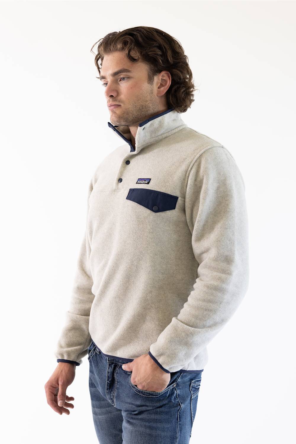 A man wearing a light beige fleece pullover sweater with a navy blue chest pocket and trim, paired with blue jeans, photographed against a plain white studio background.