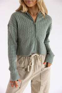 Close-up shot of a blonde woman wearing the Pink Rose cable knit hooded full-zip sweater in willow green, styled with beige drawstring pants.