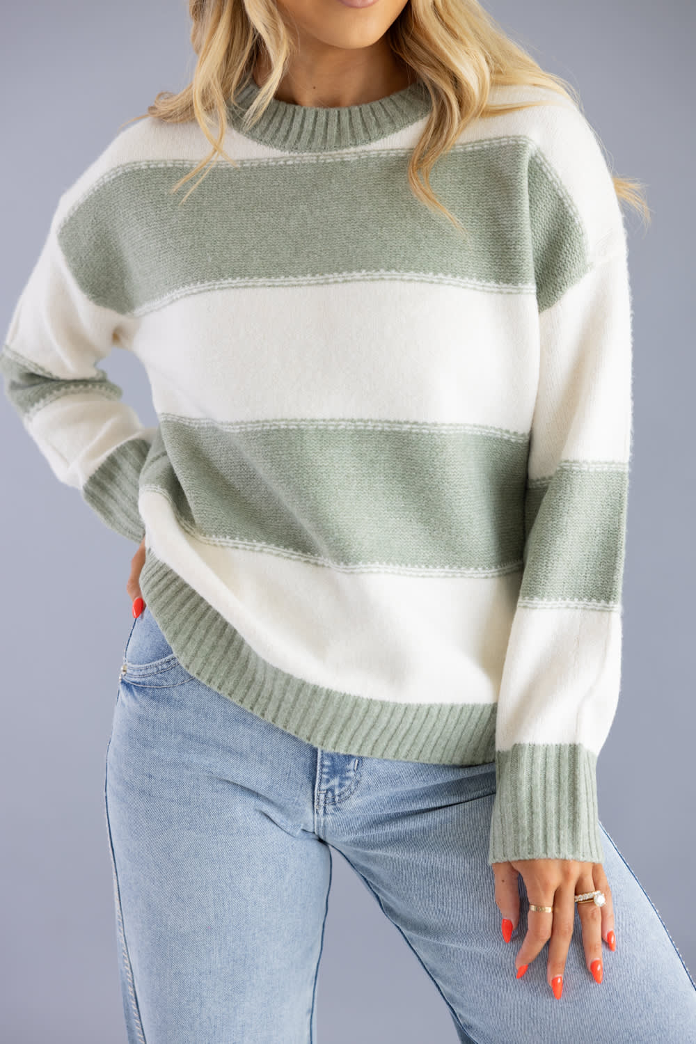Close-up shot of a woman wearing the Pink Rose cream and sage green horizontal striped crew neck sweater with ribbed collar and cuffs, paired with light-wash jeans.