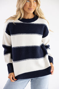 Close-up portrait view of a blonde female model wearing the Pink Rose wide-striped knit crew neck sweater in cream and navy blue, showing the collar detail and horizontal block stripe pattern.