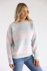 Front three-quarter view of a blonde woman wearing the Pink Rose light pink and heather grey wide-striped knit crew neck sweater, paired with blue jeans