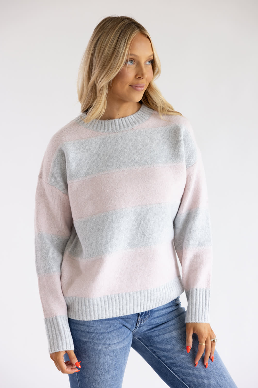 Front three-quarter view of a blonde woman wearing the Pink Rose light pink and heather grey wide-striped knit crew neck sweater, paired with blue jeans