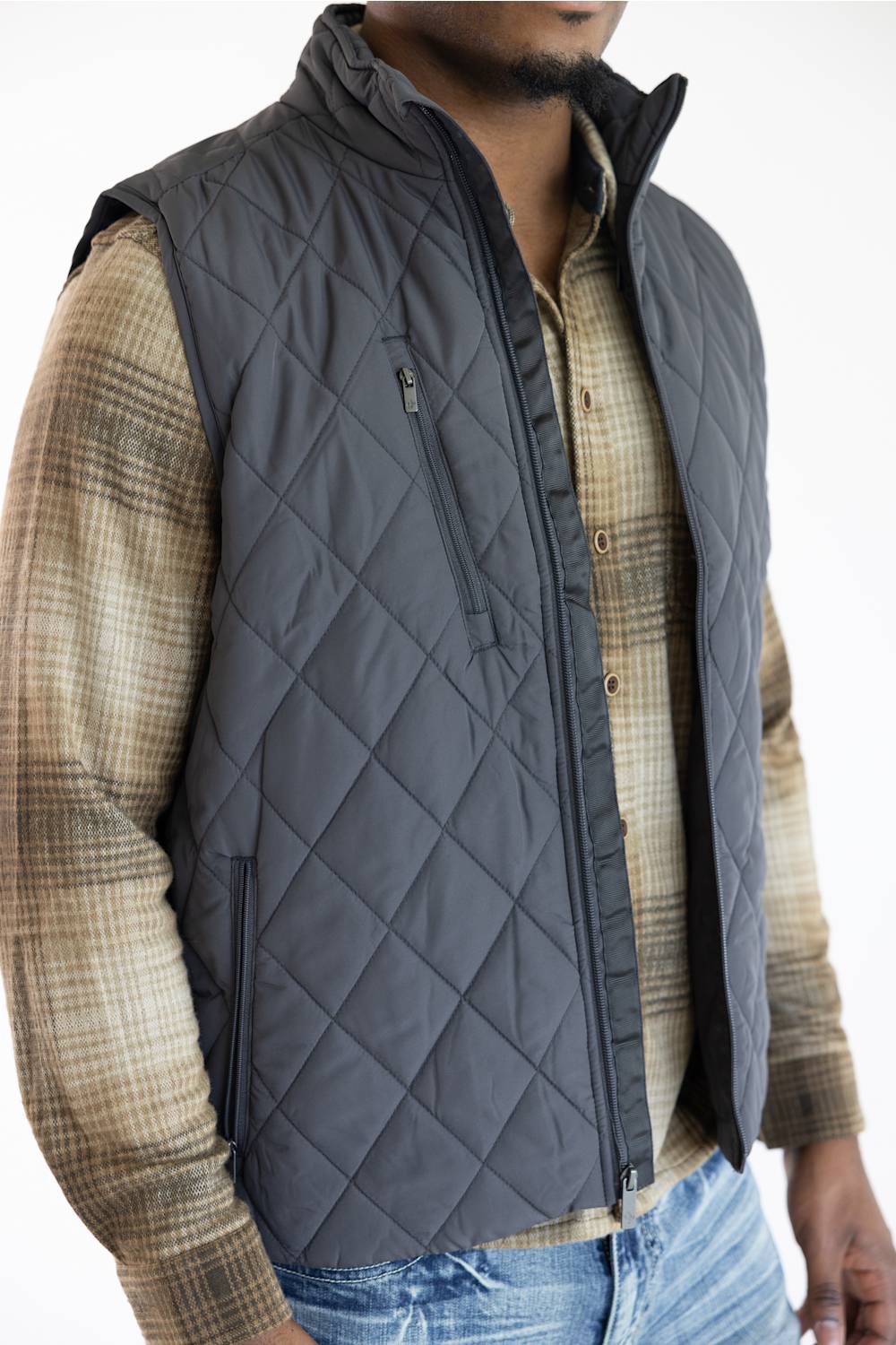 A man wearing a dark gray diamond-quilted zip-front vest over a brown and beige plaid flannel shirt, paired with blue jeans.