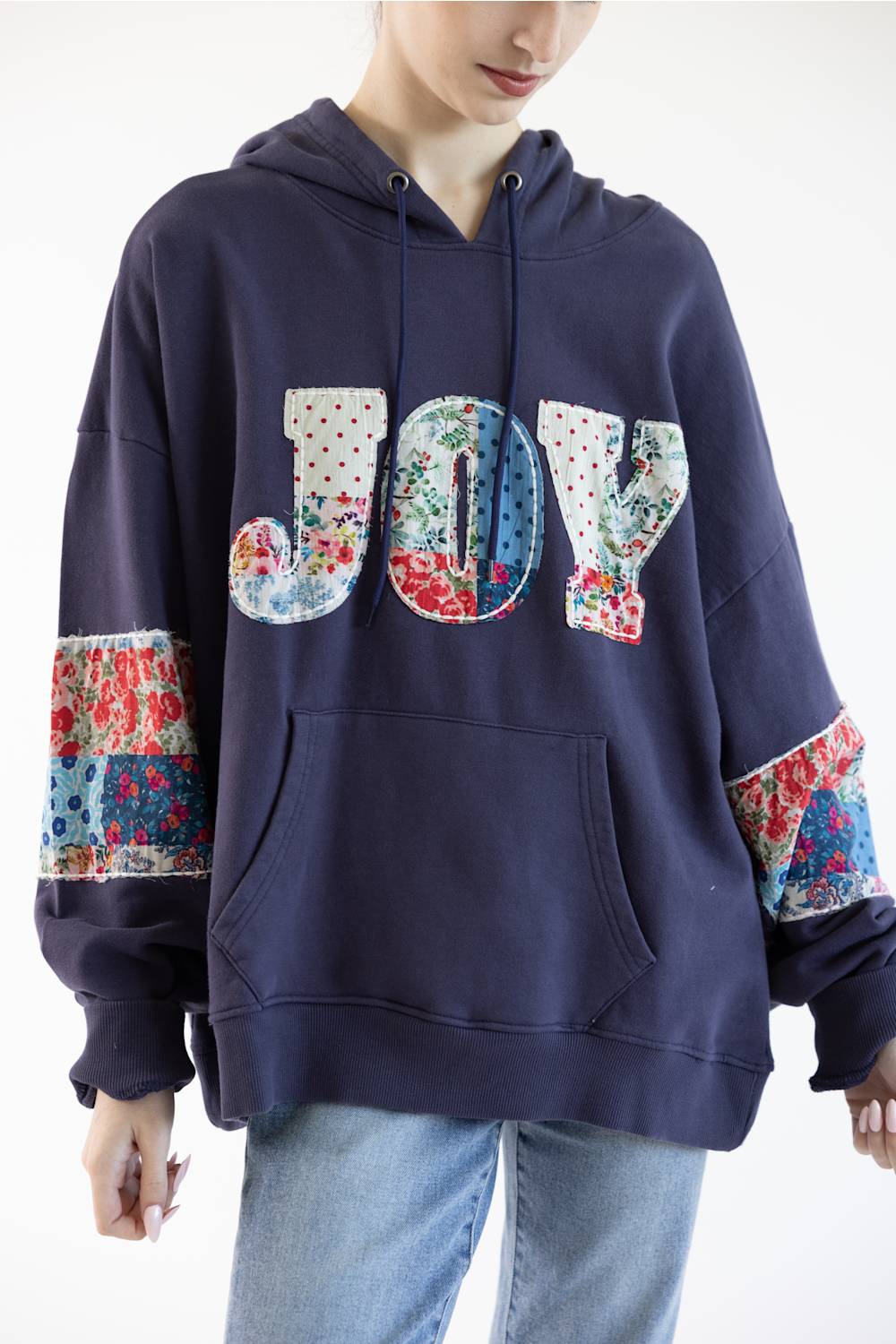 A person wearing an oversized navy hoodie with a large 'JOY' patchwork graphic across the chest and floral patchwork details on the sleeves, paired with light blue jeans against a plain white background.