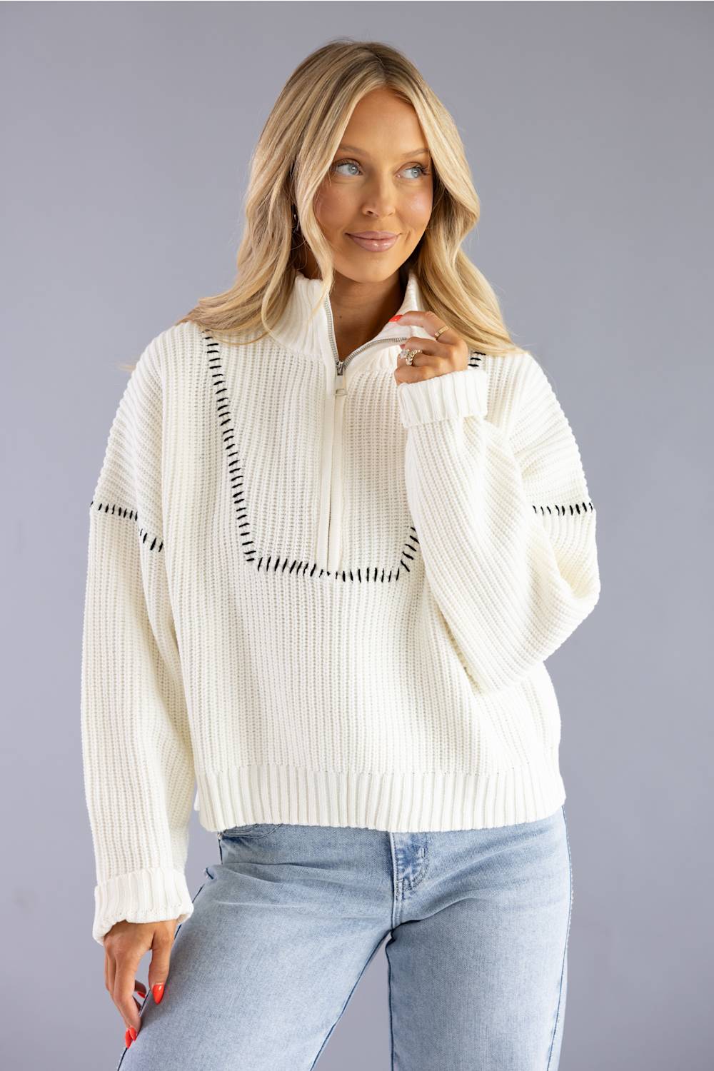 A woman wearing a cream ribbed knit half-zip sweater with black stitched geometric accents, paired with light blue jeans, against a solid gray studio background.