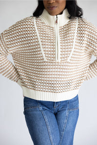 A woman wearing a cream and beige textured knit half-zip sweater with blue jeans, shown from the neck down against a light gray background.