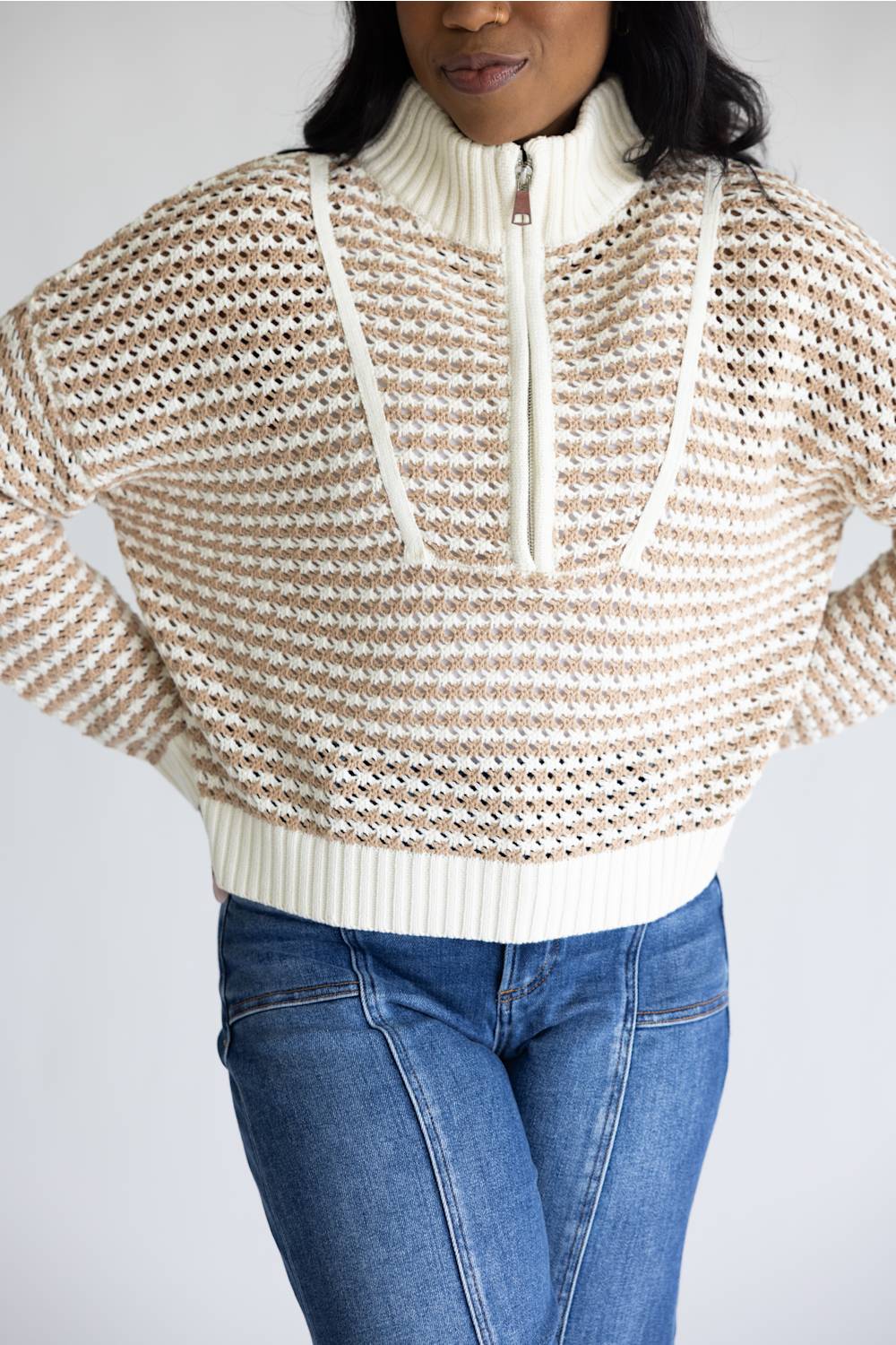 A woman wearing a cream and beige textured knit half-zip sweater with blue jeans, shown from the neck down against a light gray background.
