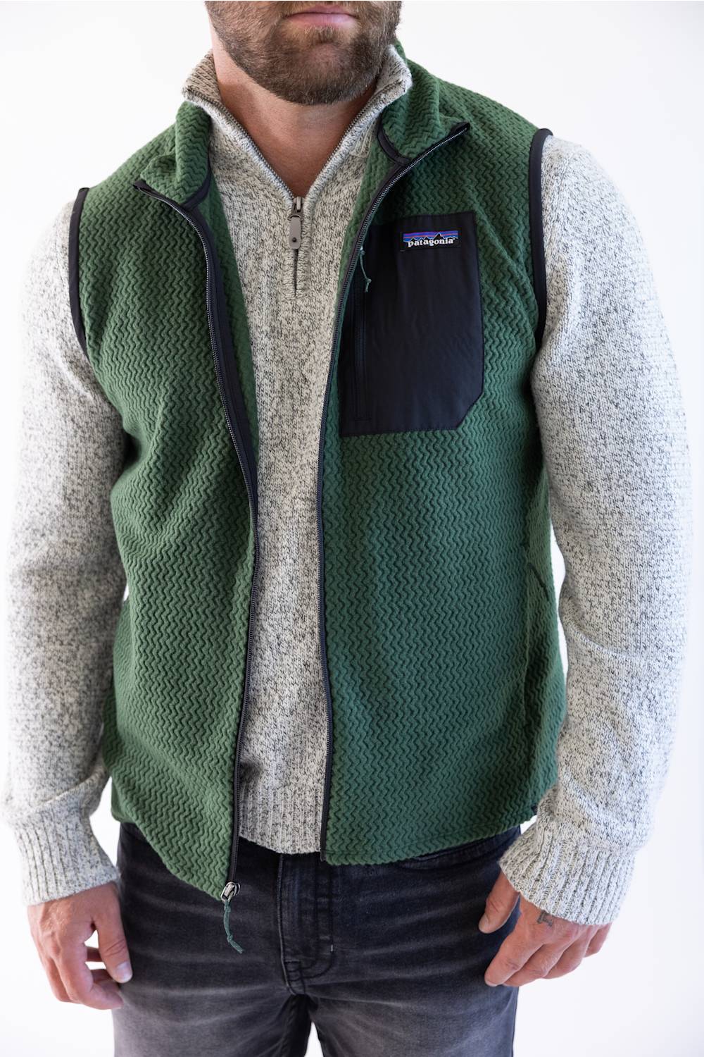 A man wearing a green Patagonia puffer vest over a beige sweater and blue jeans, shown from the chest down against a plain white background.
