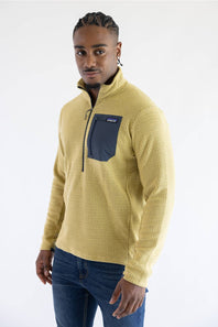 A man wearing a textured yellow quarter-zip fleece pullover with a dark navy chest pocket, paired with blue jeans, photographed against a plain white studio background.
