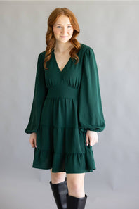 A woman modeling a dark green tiered mini dress with long balloon sleeves and black knee-high boots, standing against a neutral gray studio backdrop.