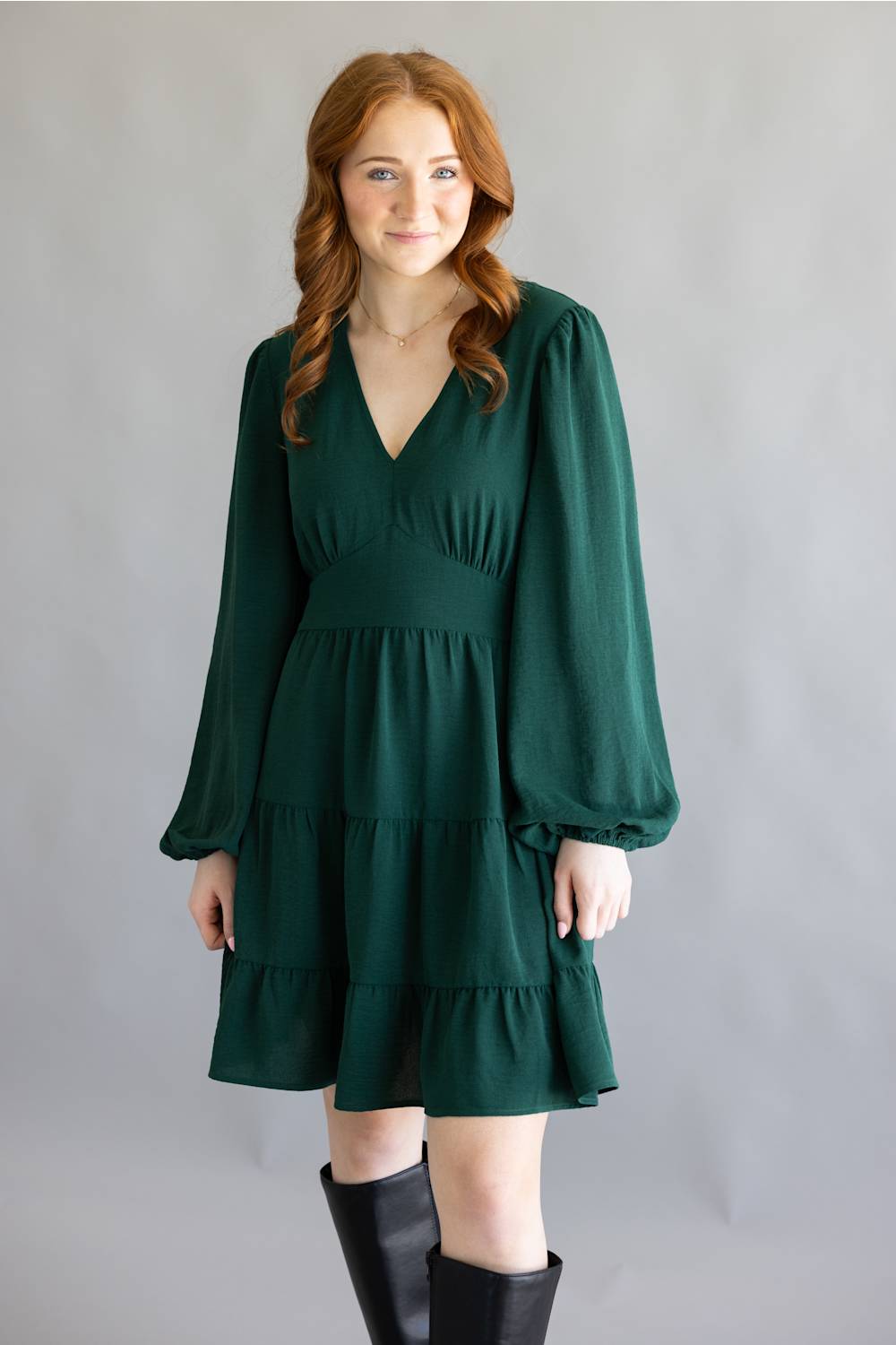 A woman modeling a dark green tiered mini dress with long balloon sleeves and black knee-high boots, standing against a neutral gray studio backdrop.