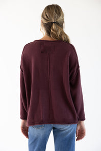 A woman viewed from the back wearing a maroon colored slouchy sweater with blue denim jeans standing in front of a white studio background
