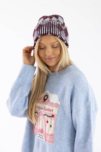 A woman wearing a light blue knit sweater with a pink Christmas market graphic and a maroon and white striped beanie, photographed against a plain white background.