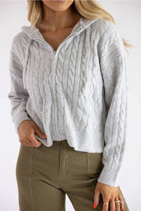 A woman wearing a light gray cable-knit zip-up hoodie sweater and olive green pants, shown from the chin down against a plain white background.