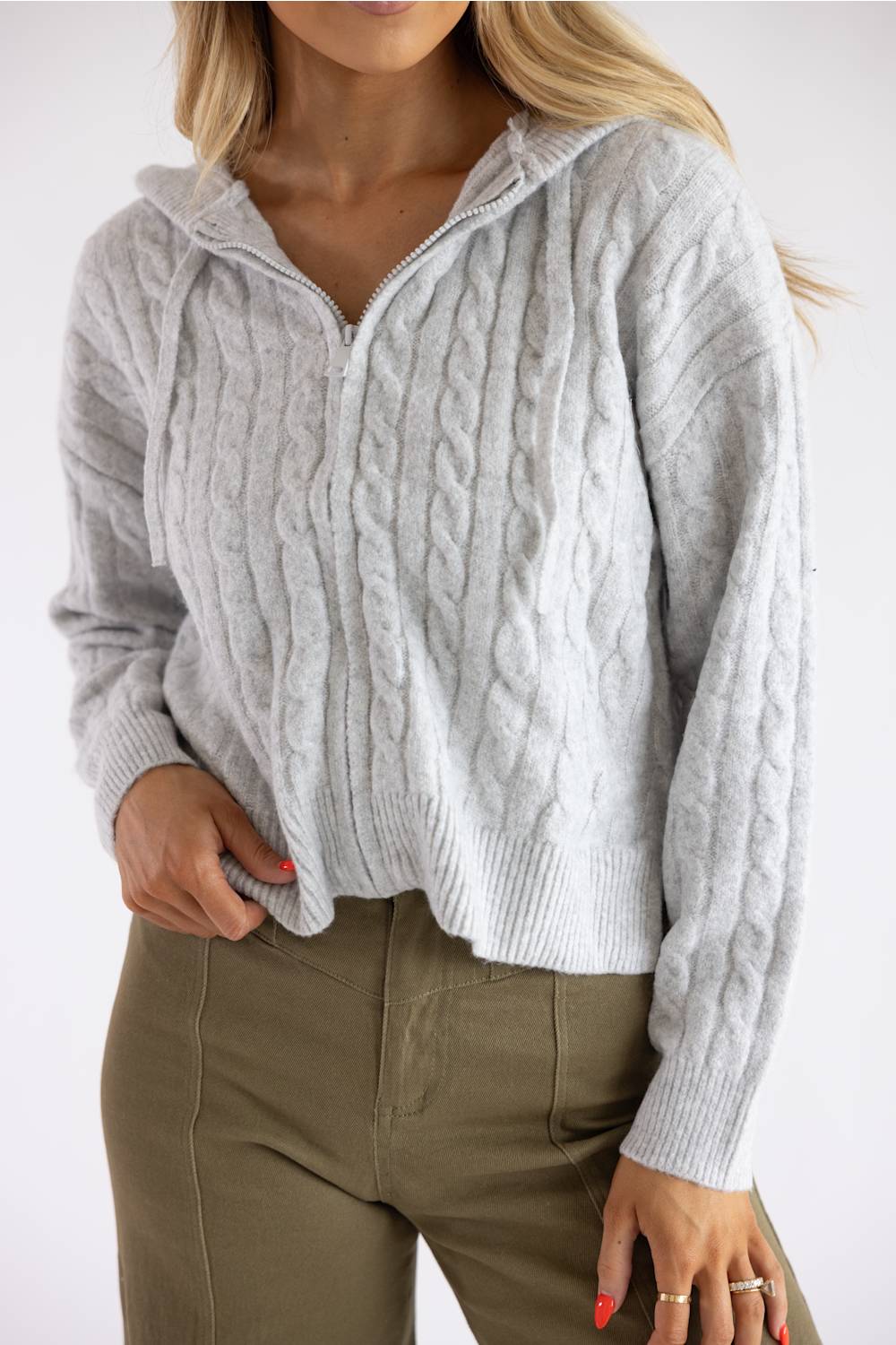A woman wearing a light gray cable-knit zip-up hoodie sweater and olive green pants, shown from the chin down against a plain white background.