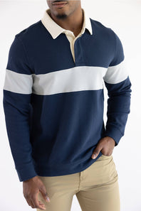 A man wearing a blue and grey color-blocked long-sleeve rugby shirt with blue jeans, standing against a plain light background.