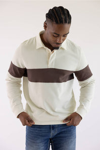 A man wearing a cream and brown color-blocked long-sleeve rugby shirt with blue jeans, standing against a plain light background.