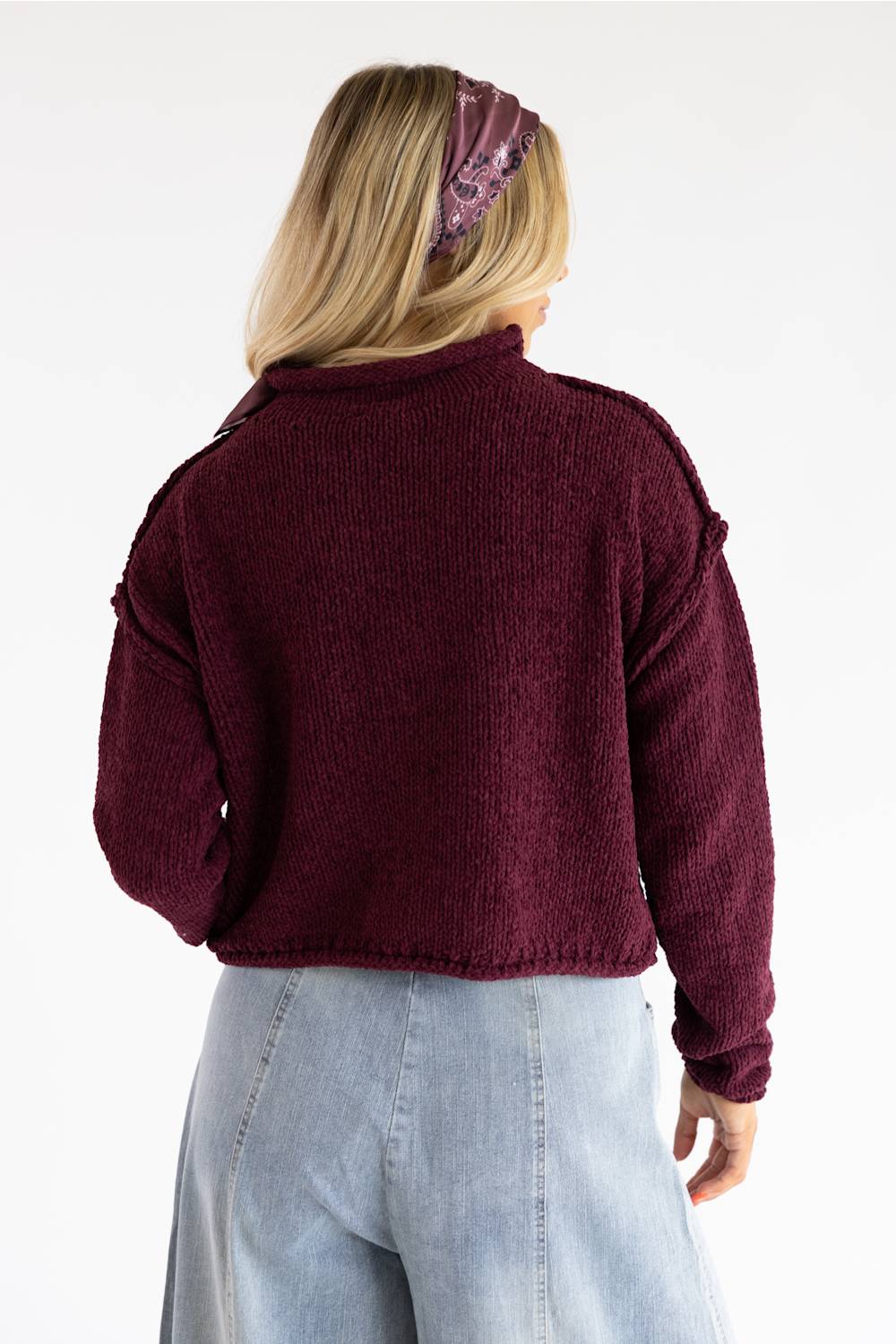 A woman shown from the back wearing a textured, cropped burgundy knit sweater and light blue jeans against a plain white background.