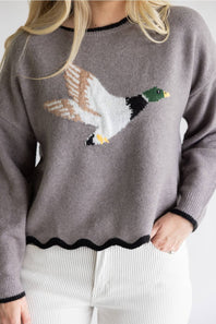 A woman wearing a gray knit sweater with a large embroidered mallard duck design across the chest, paired with white ribbed pants.