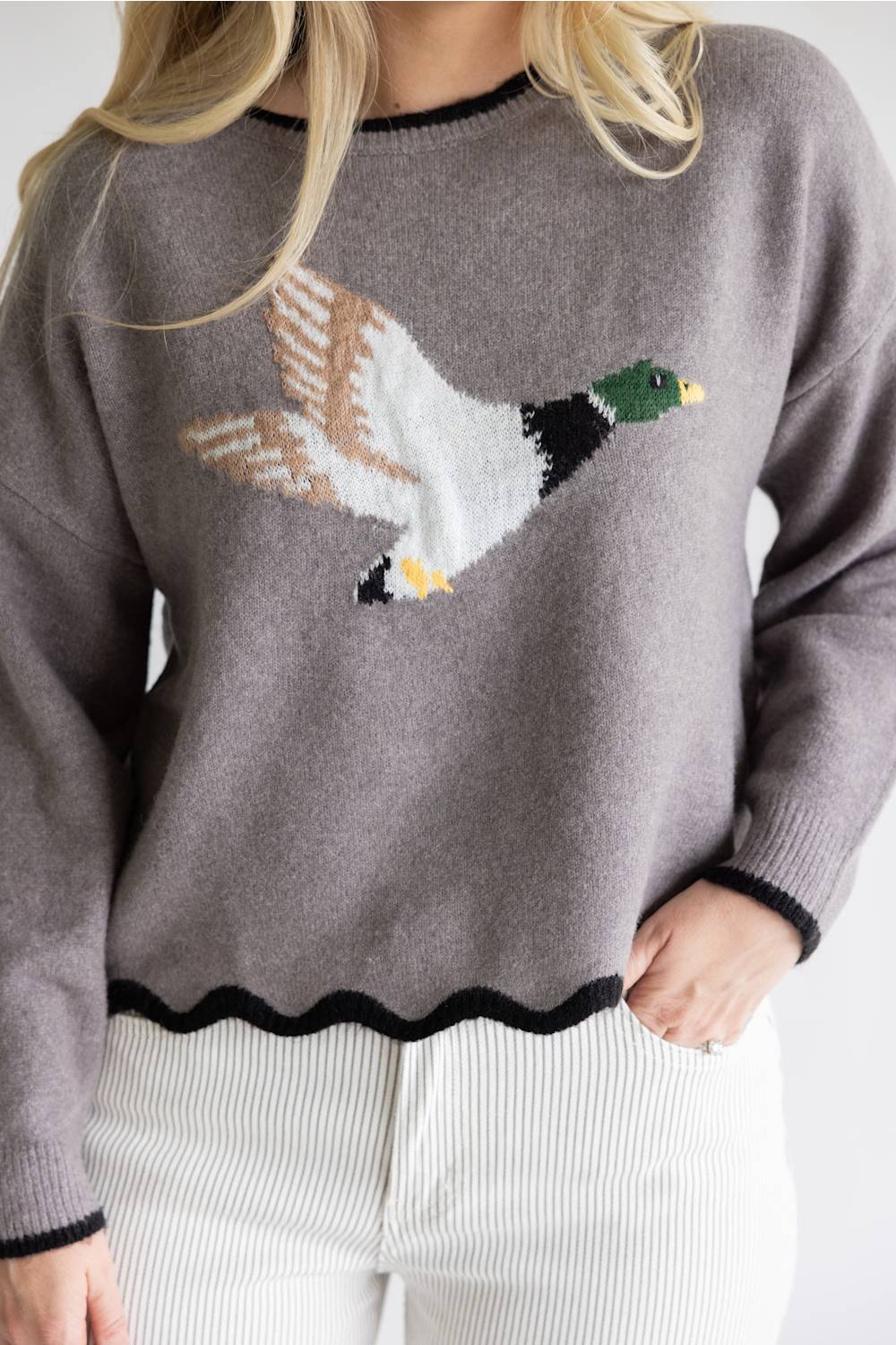 A woman wearing a gray knit sweater with a large embroidered mallard duck design across the chest, paired with white ribbed pants.