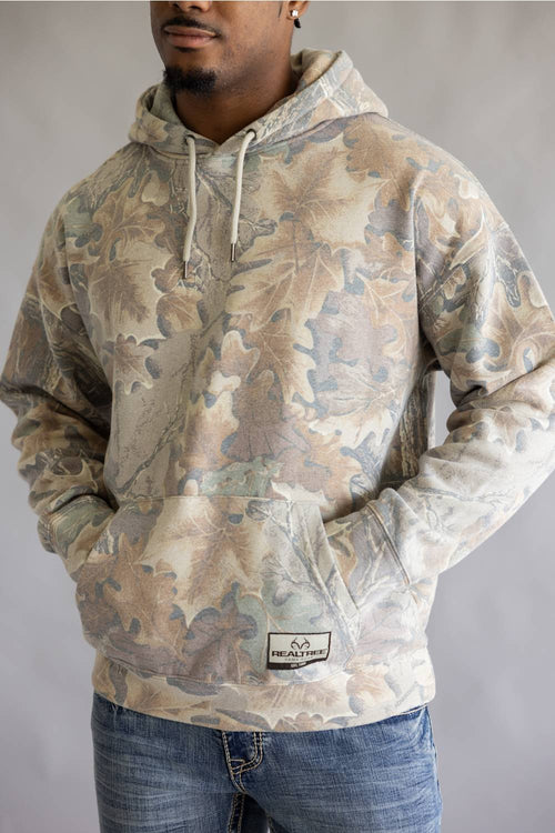 A man wearing a camouflage hoodie with an oak leaf pattern in shades of beige, brown, and gray, paired with blue jeans against a neutral gray background.