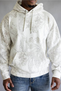 A man wearing a white and gray camo pattern hoodie with blue jeans, standing against a neutral gray studio background.