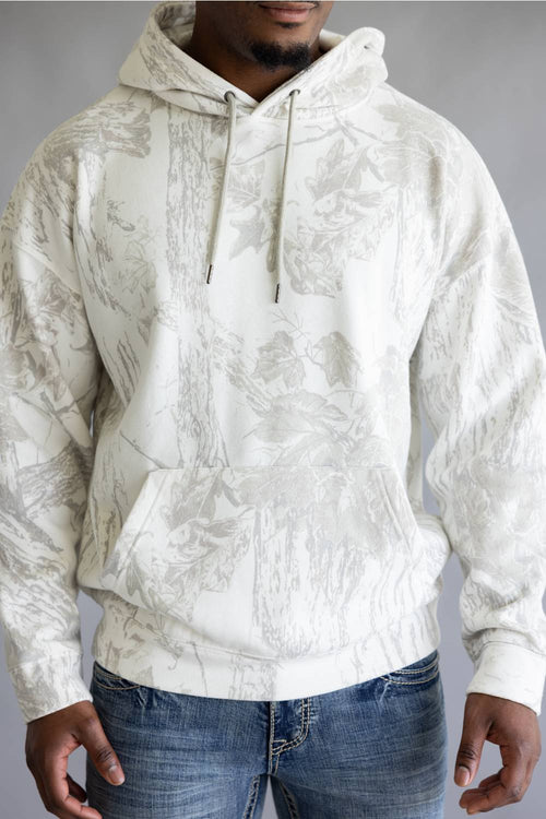 A man wearing a white and gray camo pattern hoodie with blue jeans, standing against a neutral gray studio background.