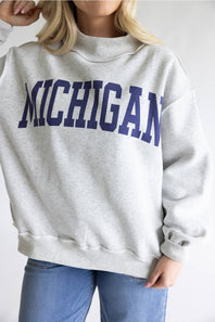 A woman wearing a light heather gray sweatshirt with 'MICHIGAN' printed across the chest in dark navy blue collegiate lettering, paired with blue jeans.